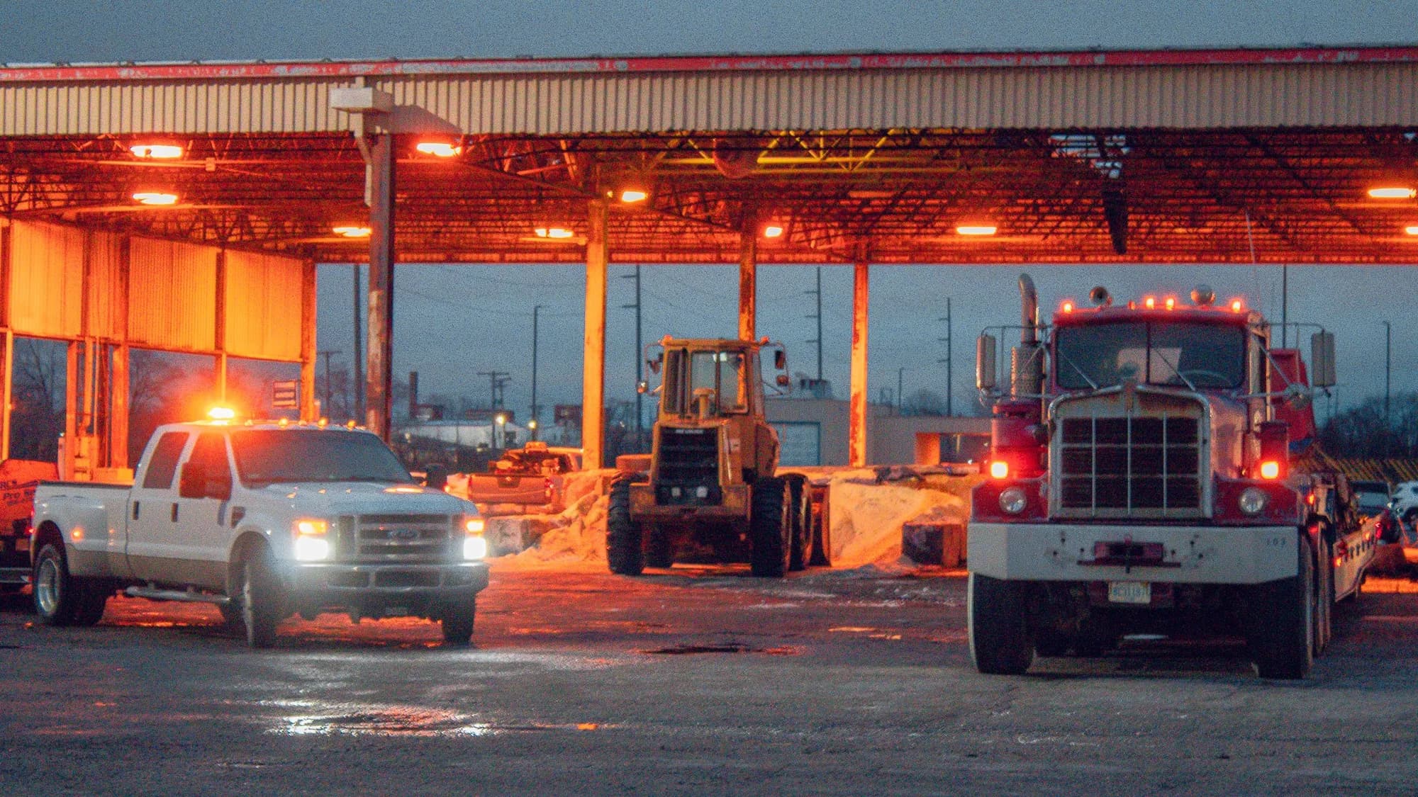 Pickups, a loader and a semi truck staged under a lit canopy at night
