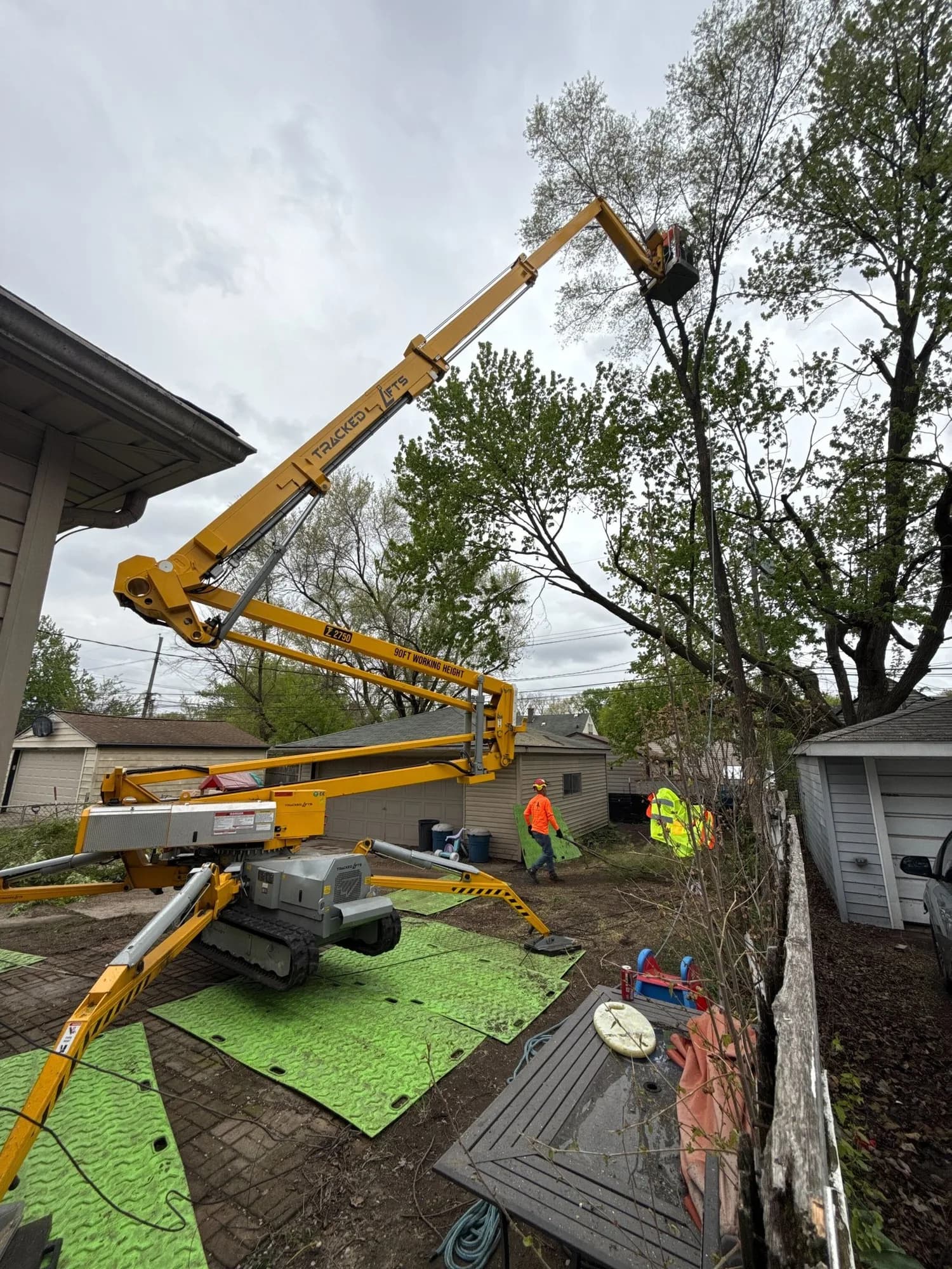 Tracked spider lift set up on protection mats in a narrow backyard, reaching into a tree