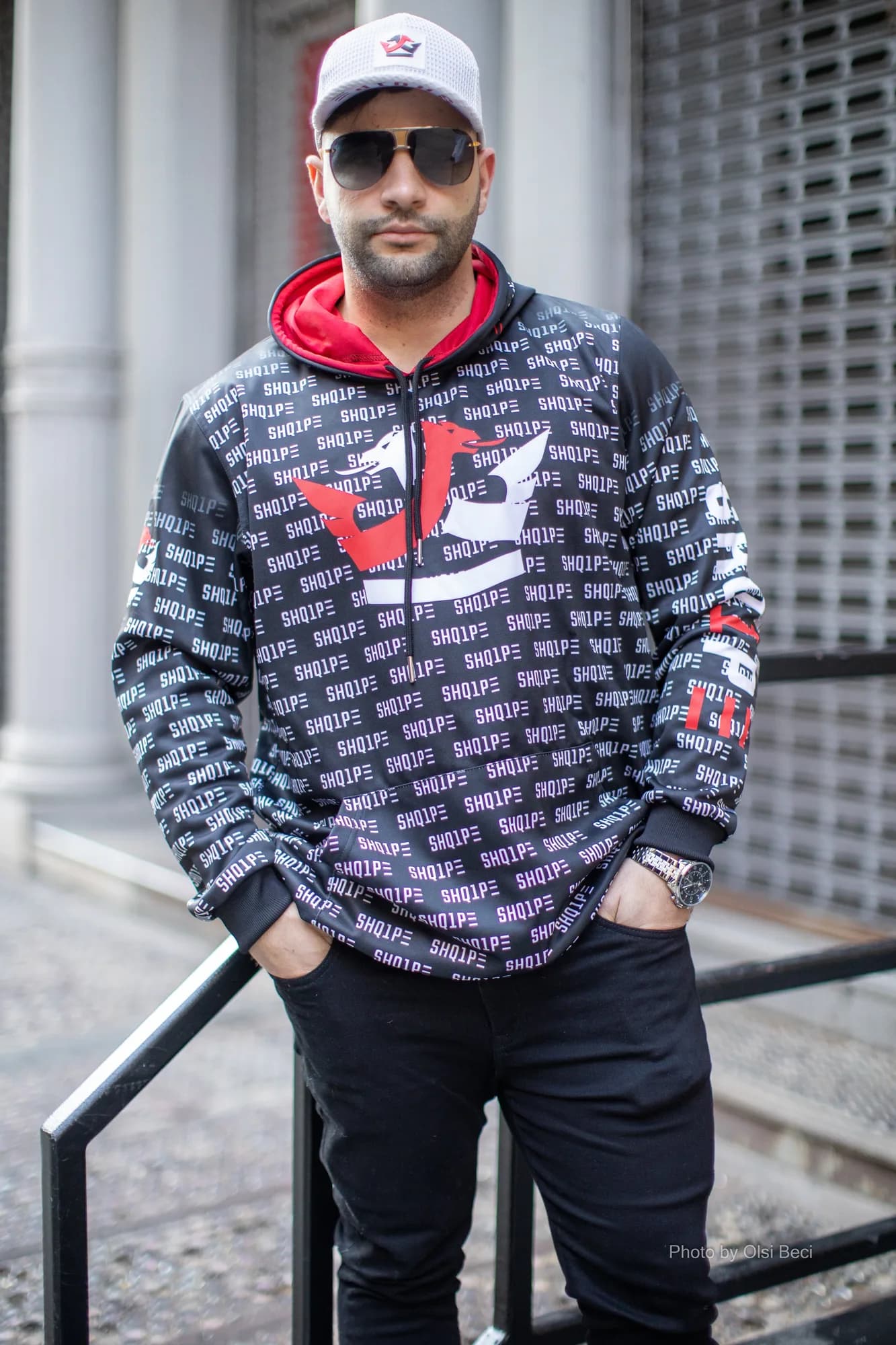 Man in a black LEK all-over print hoodie and white cap on a city street